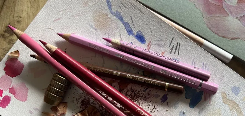 Pink and red coloured pencils, shavings and a brass sharpener on a watercolour-spattered sheet.